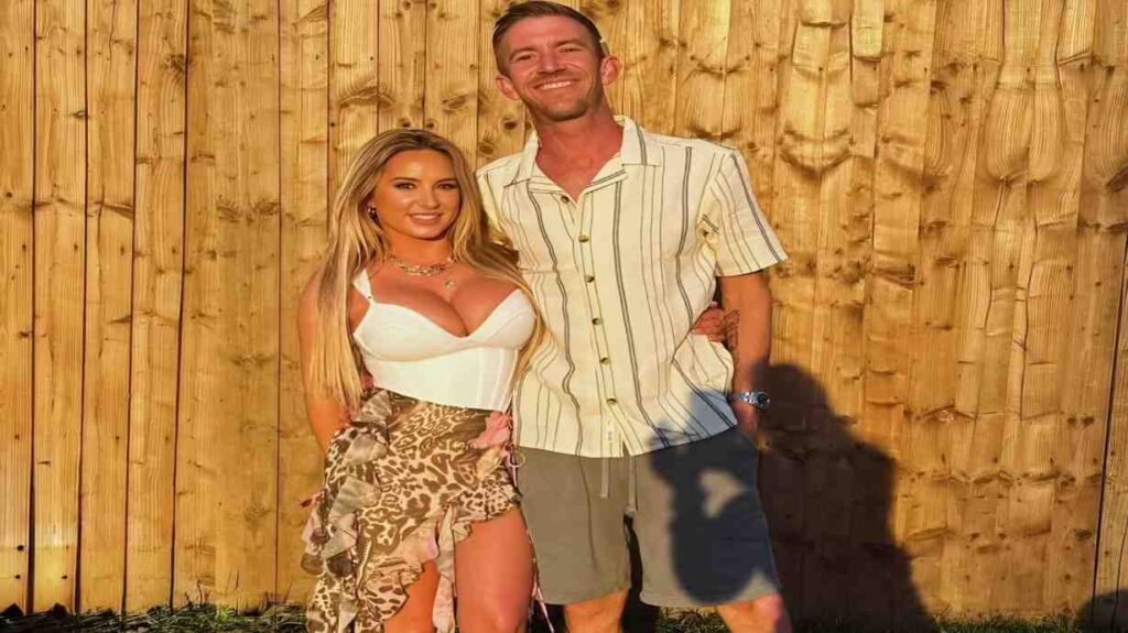 Danny D and woman stand side by side in front of a wooden fence, smiling and dressed casually