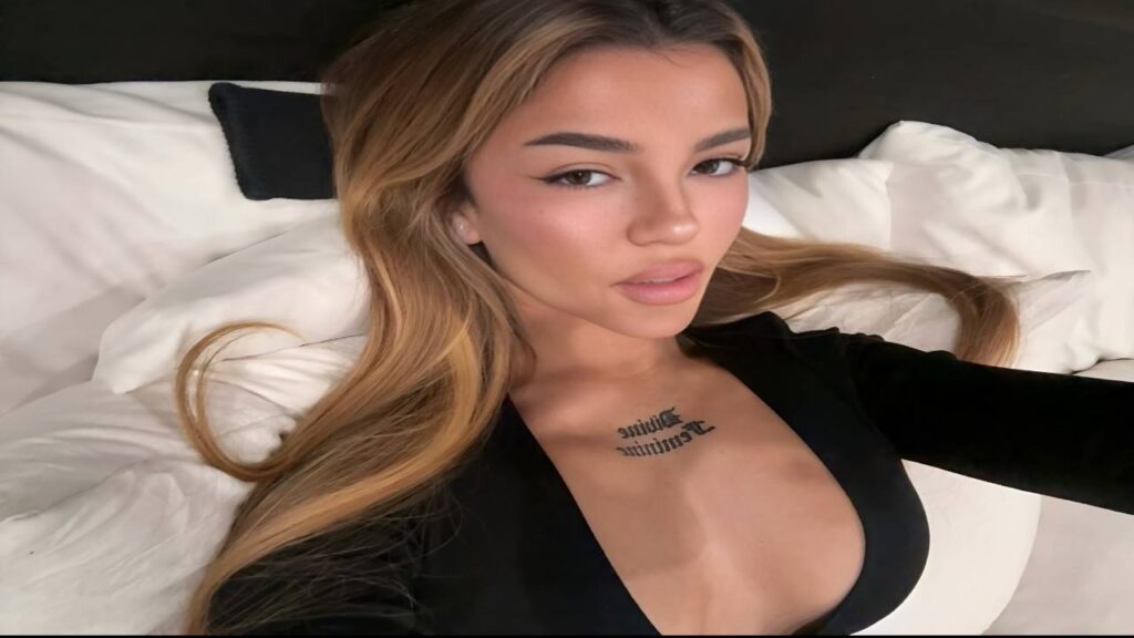 Ellie Luna in a black top and bra reclines on a bed, appearing relaxed and contemplative.