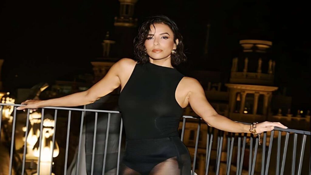 Eva Longoria in a black top and stockings poses confidently on a balcony, showcasing a stylish and elegant look