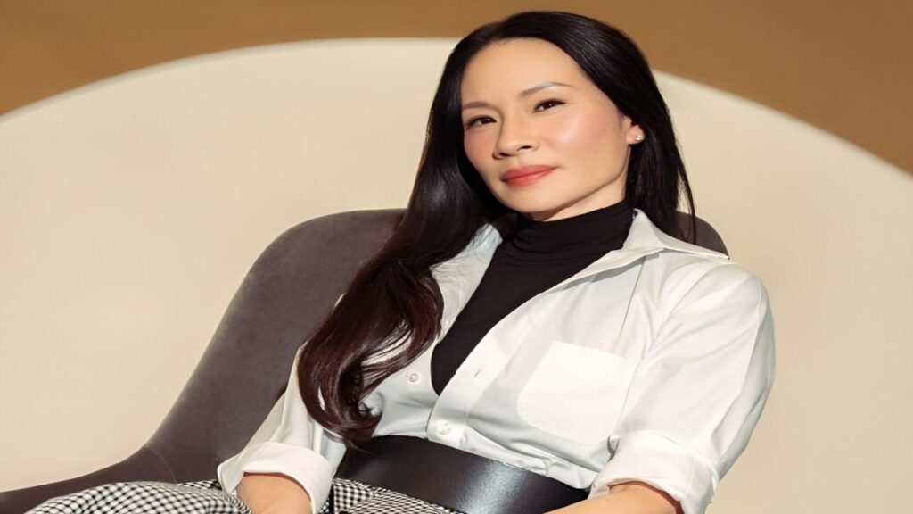 Lucy Liu sitting in a chair with her legs crossed, looking thoughtfully to the side.