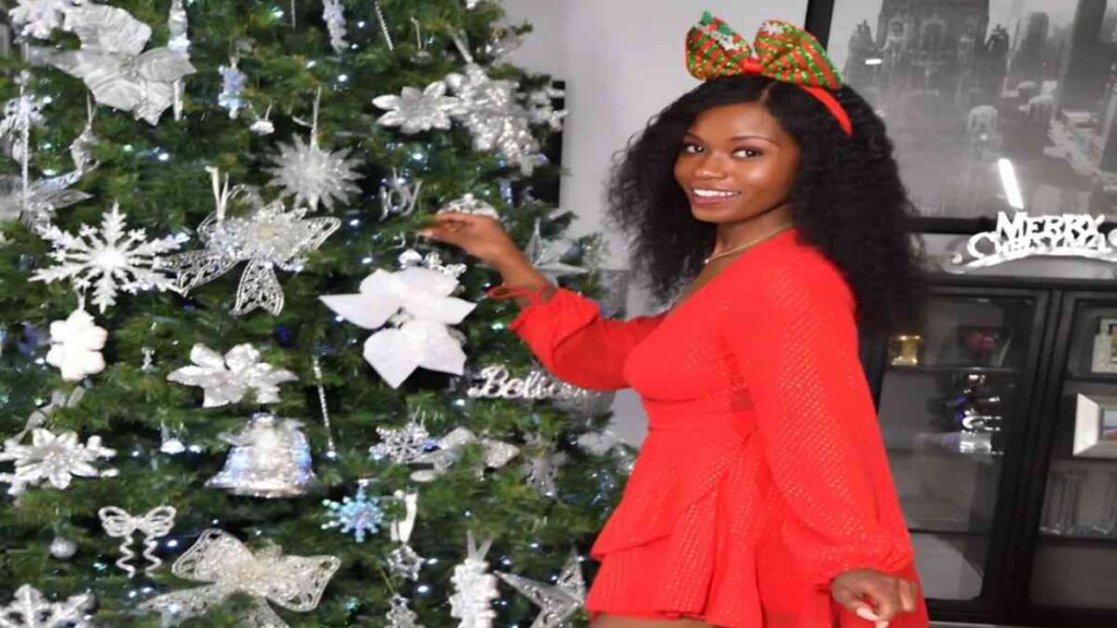 Jezabel Vessir in a red dress stands beside a decorated Christmas tree, creating a festive holiday atmosphere