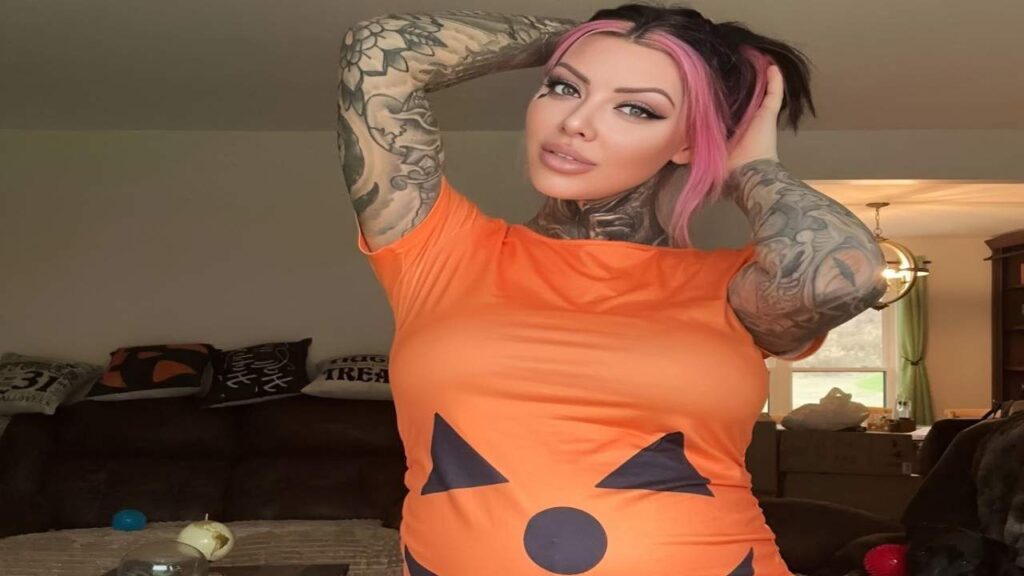 Karma Rx with tattoos wearing a vibrant pumpkin costume, smiling and posing for the camera in a festive setting