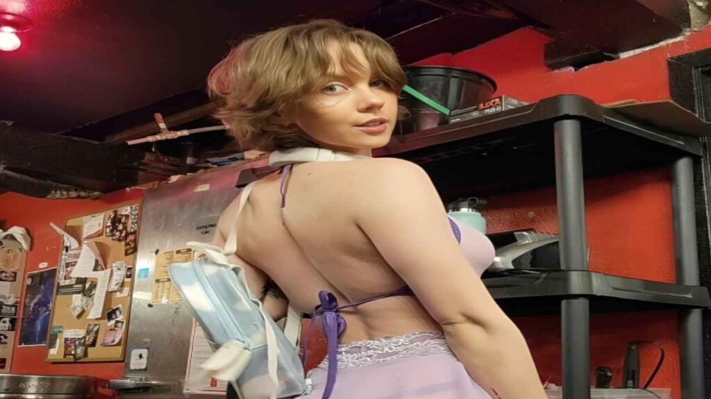 Kayla Coyote in a purple dress poses confidently in a modern kitchen, showcasing her stylish outfit and the kitchen's decor.