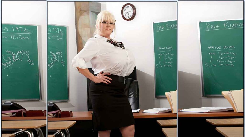 Kayla Kleevage in a skirt and blouse stands in front of a blackboard, appearing engaged in teaching or presenting information
