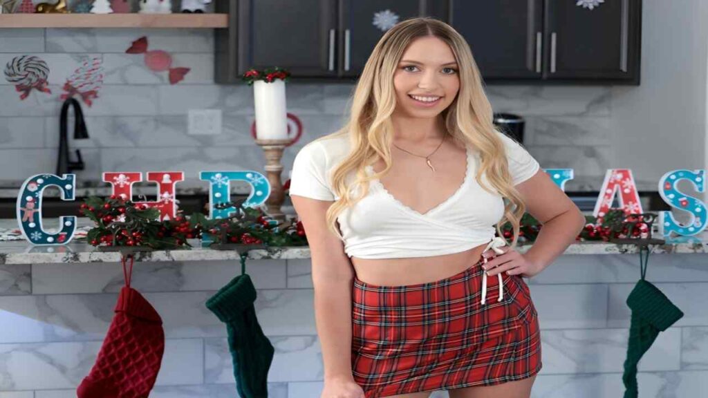 Khloe Kapri in a plaid skirt and white top stands smiling in front of a decorated Christmas tree.