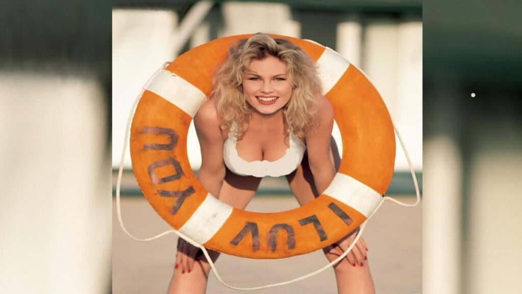 Kimberly McArthur in a bikini poses confidently with a life preserver, showcasing a vibrant beach setting.