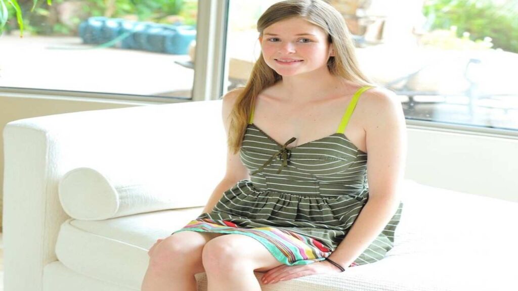 Lara Brookes in a striped dress sits comfortably on a couch, smiling and relaxed in a cozy living room setting