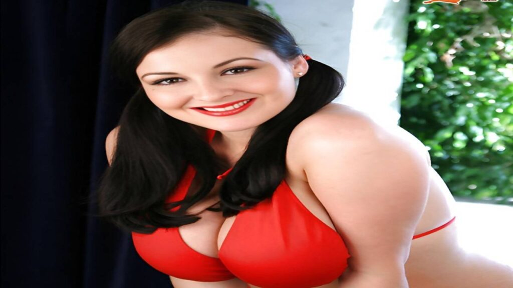 Lorna Morgan poses confidently in a red bikini, smiling for the camera against a sunny beach backdrop