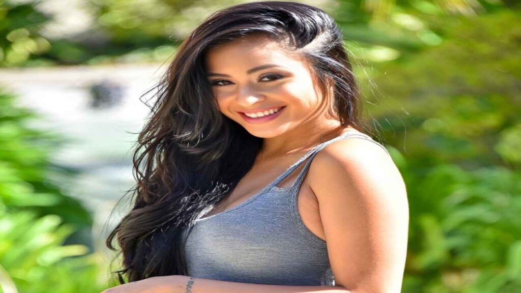 Michelle Maylene with long black hair wearing a gray tank top stands confidently, showcasing her style and poise.