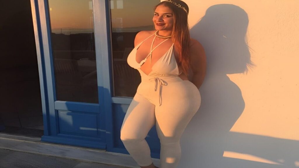 Miss Raquel in a white jumpsuit confidently poses for the camera with a smile