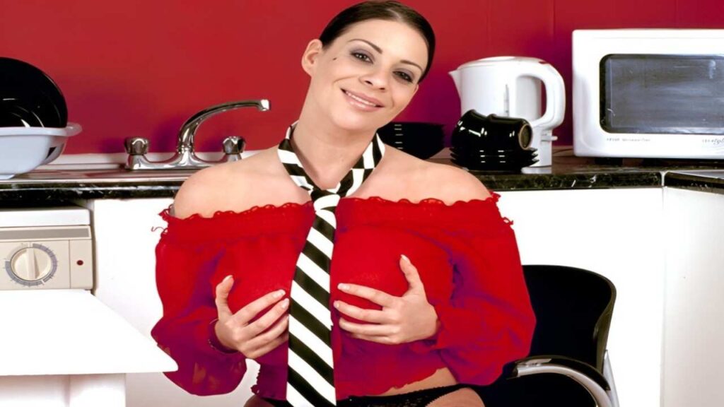 Nadine Jansen in a red shirt and tie stands in front of a stove, preparing a meal with a focused expression