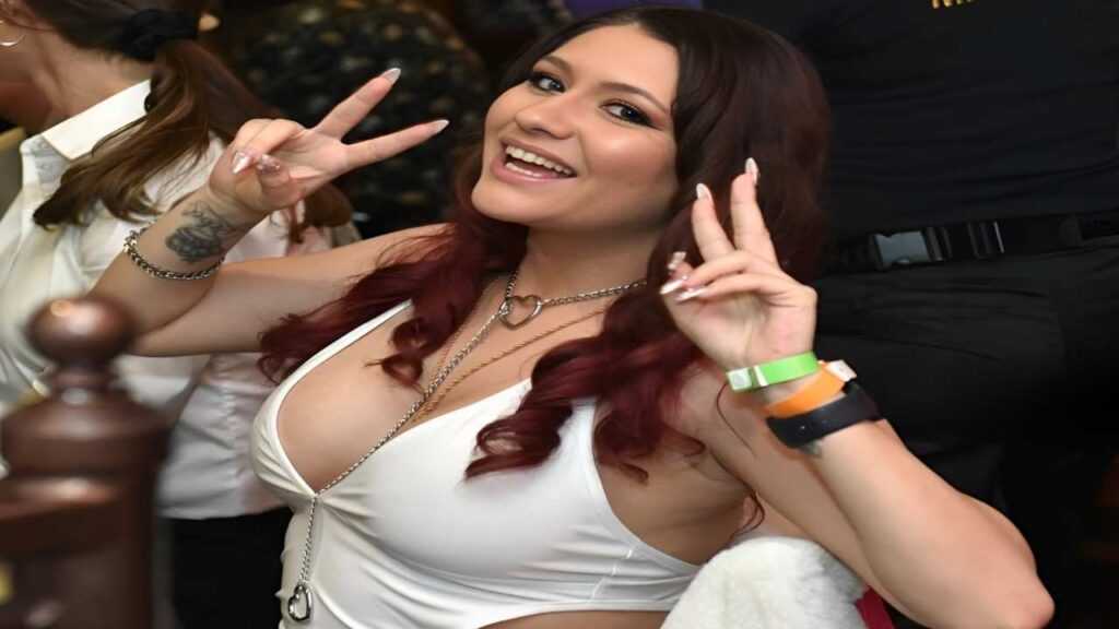Rebel Lynn with red hair in a white top is smiling and making a peace sign with her right hand.
