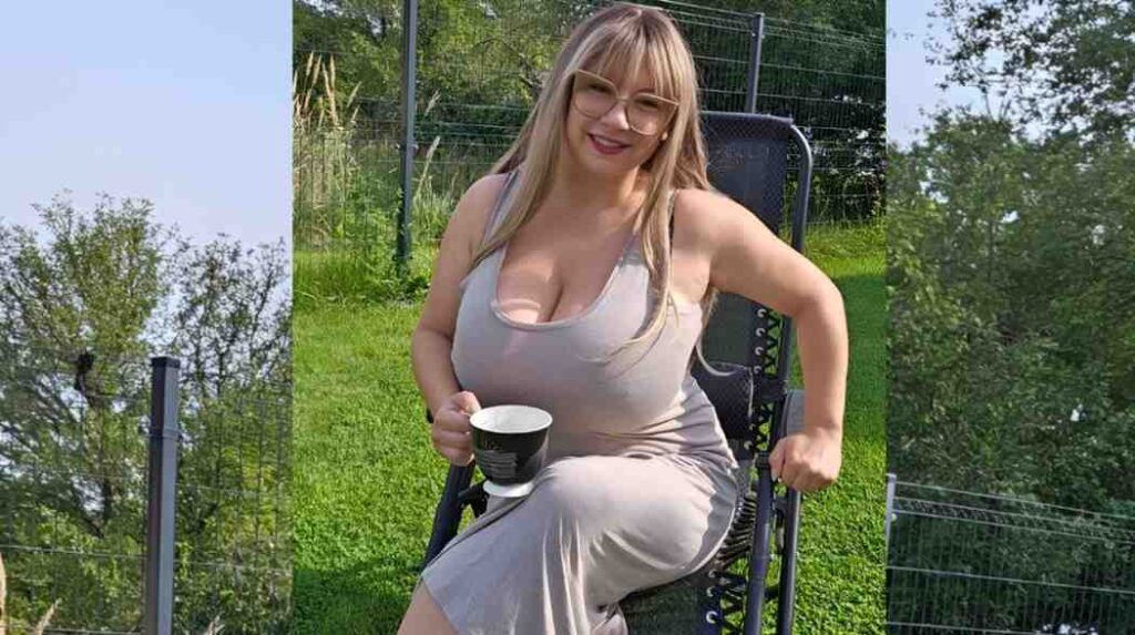 Samanta Lily in a dress sits on a chair, holding a cup of coffee, with a relaxed expression.