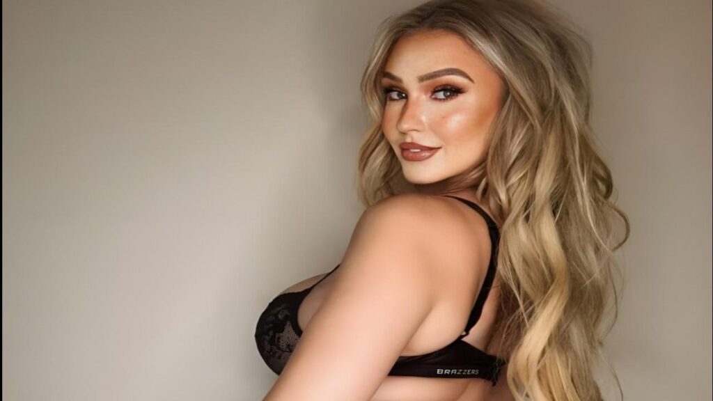 Sophie Reade in black bra, long wavy hair, sultry makeup, posing sideways against a plain beige wall.