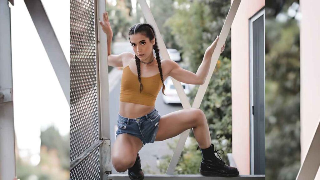 Abbie Maley with two braids in a mustard crop top and denim shorts crouches on a metal frame, gripping rails outdoors.