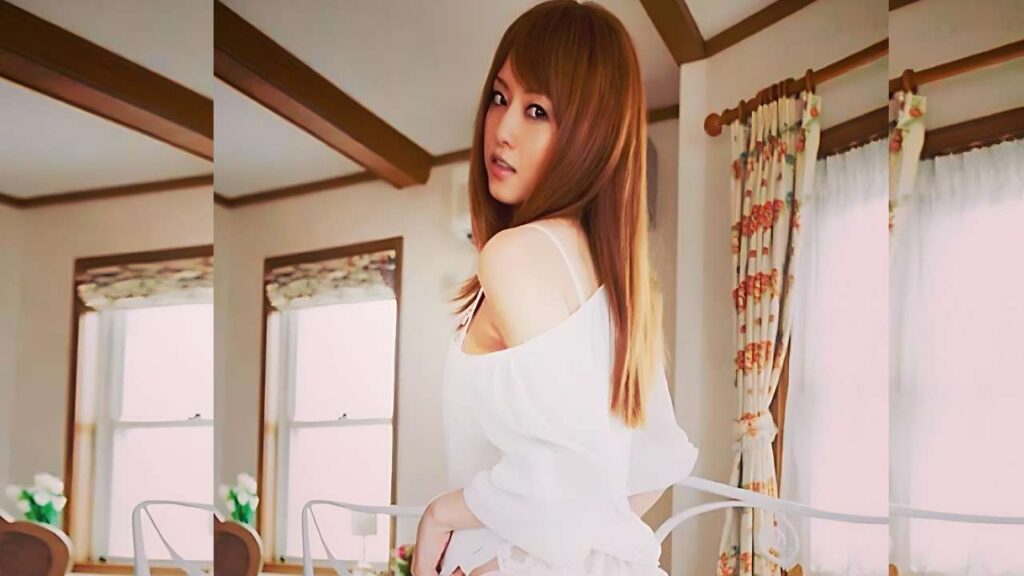 Akiho Yoshizawa (吉沢明歩 ) with long brown hair in a white off-shoulder top poses in a room with beams and floral curtains, looking at camera