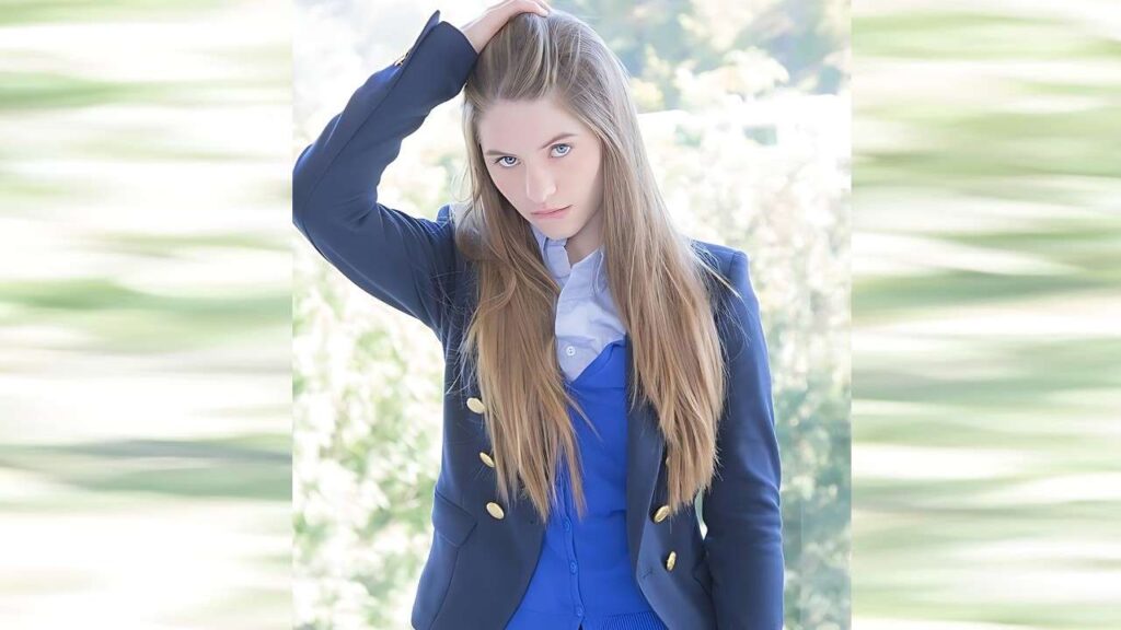 Alice March with long blonde hair in a navy blazer with gold buttons, blue top, posing with hand on her head outdoors.