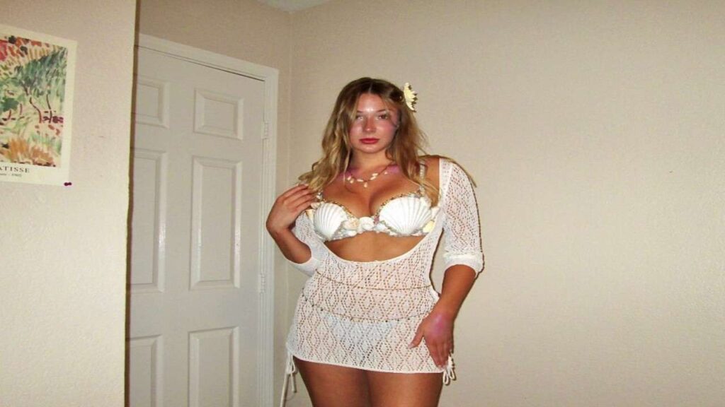 Amanda Lane in a white crocheted cover-up over a seashell bikini poses in a beige room, with a flower in her hair.