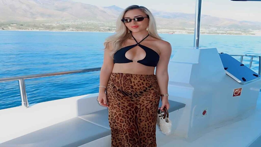 Amy Moore in a black halter bikini top and leopard pants stands on a white yacht deck holding a woven bag, blue sea behind
