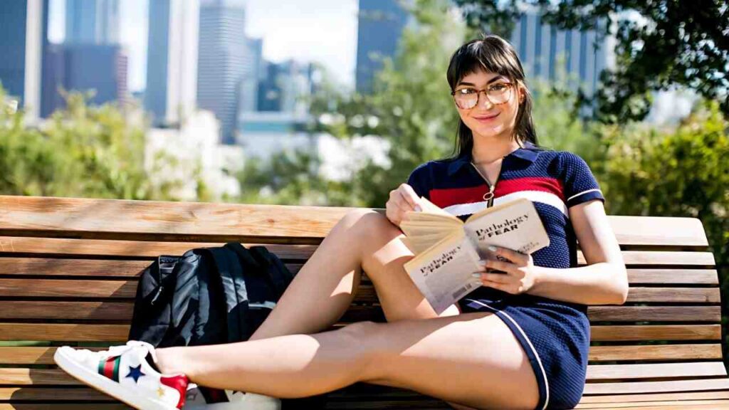 Ana Rose in a navy polo with red stripes sits on a bench, reading a book beside a backpack, city skyline behind.