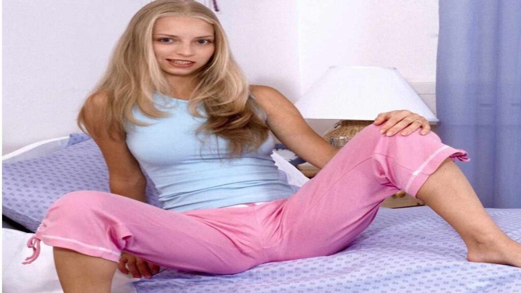 Ashley Lightspeed lounging on pastel bed, wearing a light blue sleeveless top and pink pajama pants, with one leg extended.