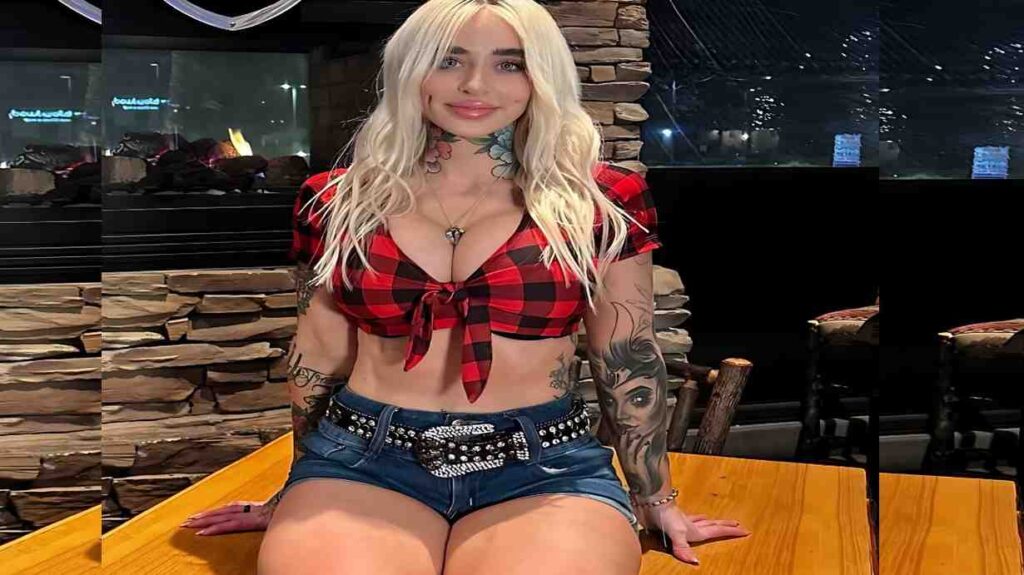 Ashley Wolf with long hair sits at a wooden table, wearing a red plaid tie-front top and denim shorts; tattooed arms.