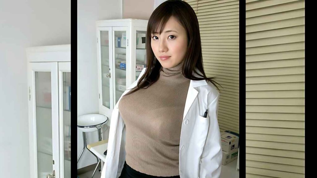 Azusa Nagasawa in a white lab coat over a beige turtleneck smiles gently in clinical room, with glass cabinets and blinds behind.