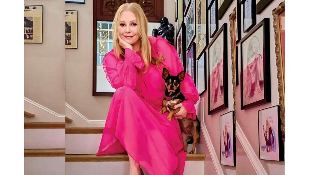 Bebe Buell in a bright pink flowing dress sits on wooden stairs, holding a small Chihuahua, while a wall of framed art lines the hallway.