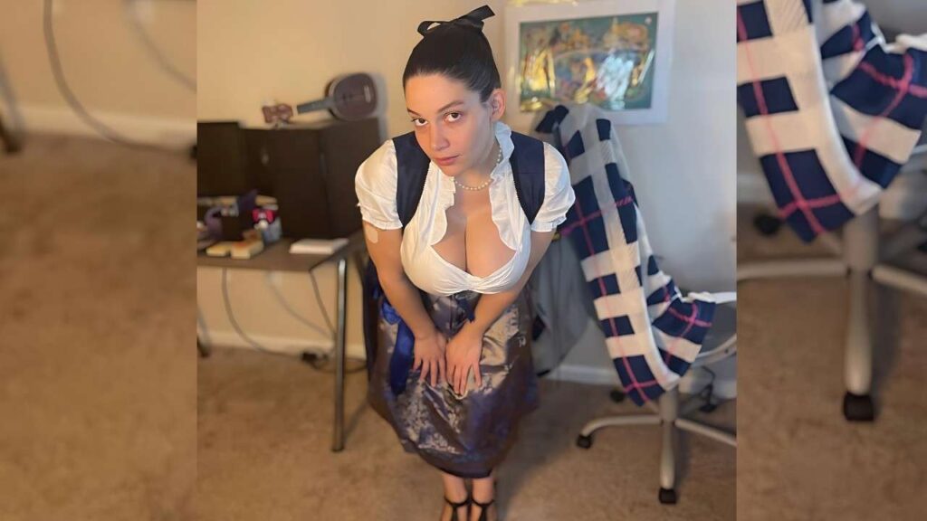 Brooklyn Springvalley in white blouse and dirndl leans forward, hands on knees, backpack on shoulders, hair in a bow, in a cluttered room.