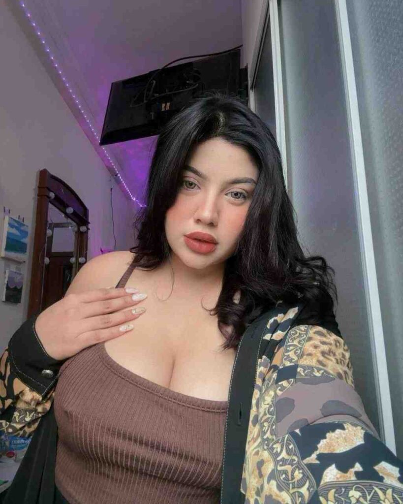 Dea Ayu with dark hair, bold makeup, wearing a brown ribbed top and patterned jacket, standing by a window.