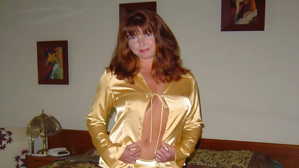 Dee Delmar in a shimmering gold satin blouse, tied at the bust, posing by a bed with a lamp and framed art.