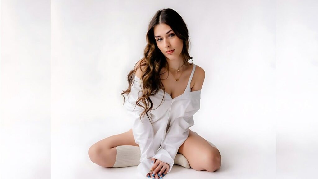 Erica Fontes sits on a white studio floor in an oversized white shirt, knee-high socks, and necklace, with long wavy hair.