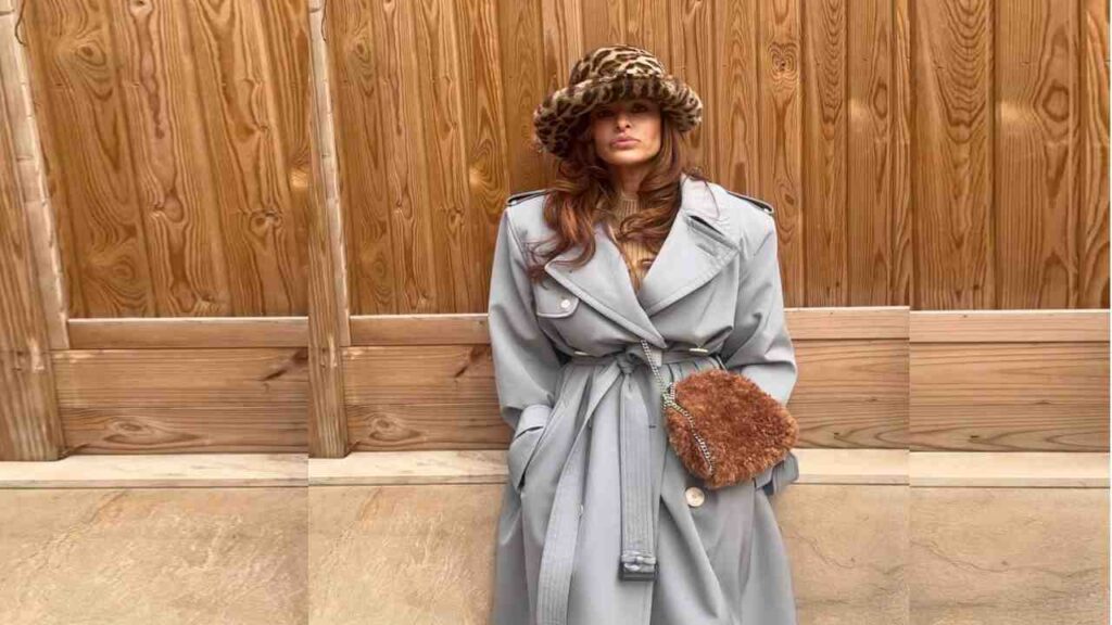 Eva Mendes in a pale blue belted trench coat, leopard-print bucket hat, and brown fuzy handbag, standing before a wooden fence.