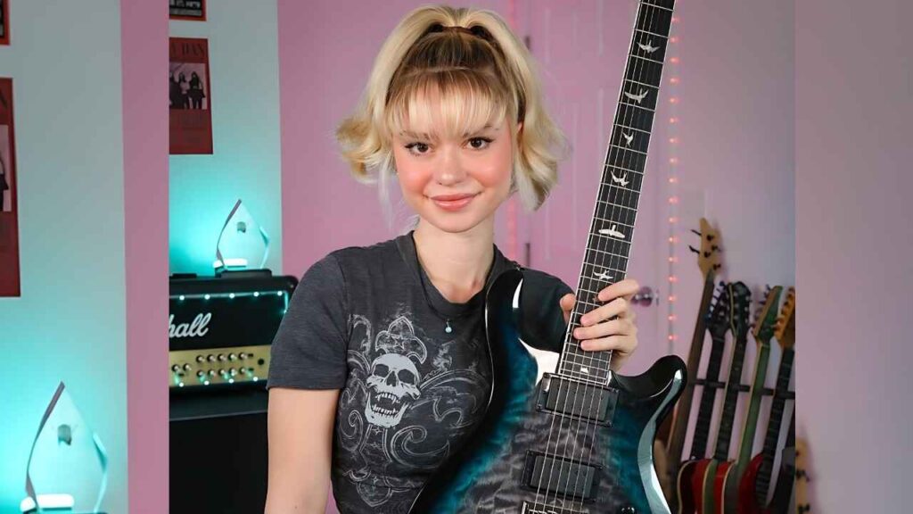 Lexi Rose with a blonde high ponytail holds a teal-black electric guitar, standing in a pastel pink music room with a Marshall amp behind and several guitars on stands; she wears a dark skull-graphic tee and the room is lit by teal string lights.
