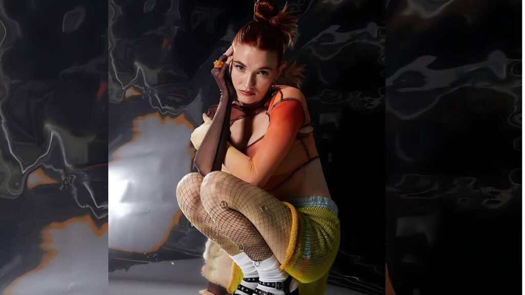 Mandy Lee crouches in orange top, fishnet tights, and a yellow skirt, chin resting on gloved hand before dark backdrop.