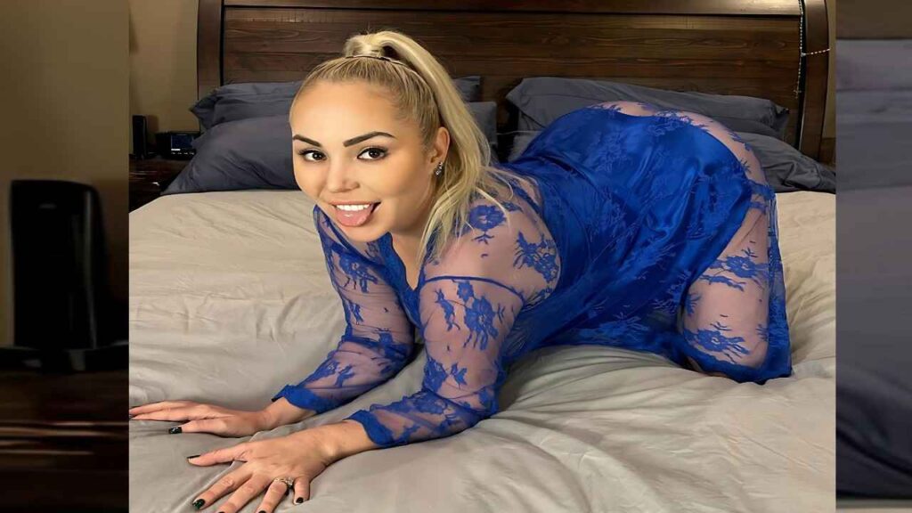 Mary Carey with ponytail wearing sheer blue lace dress on hands and knees on bed, tongue out, gray sheets and headboard.