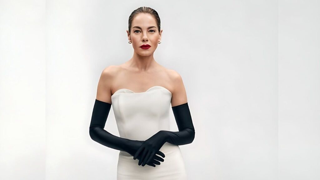 Michelle Monaghan in a strapless white gown and long black gloves stands with hands crossed at the waist, against a pale backdrop