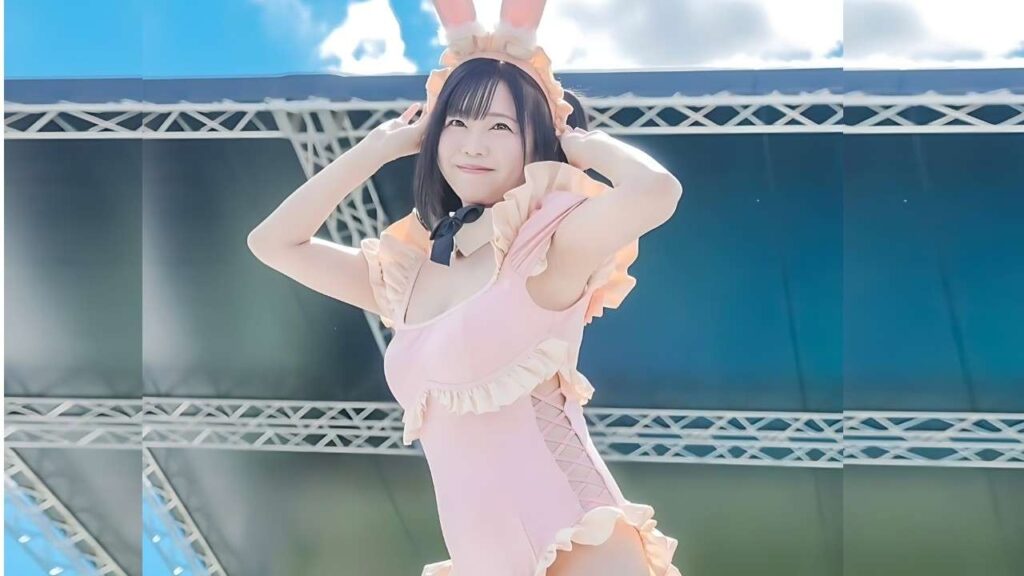 Miharu Usa in a pastel pink bunny outfit with ears and ruffles, smiling and posing with hands near ears on an outdoor stage.