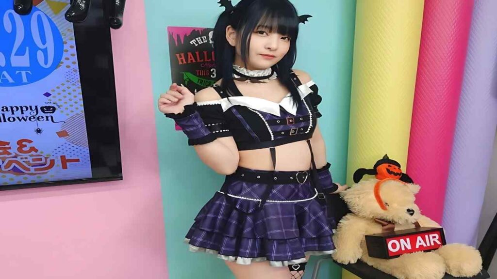 Mikako Abe ( あべみかこ ) in purple plaid outfit with bat-ear headband poses in a pastel studio, Halloween poster behind, a teddy bear with ON AIR sign nearby.