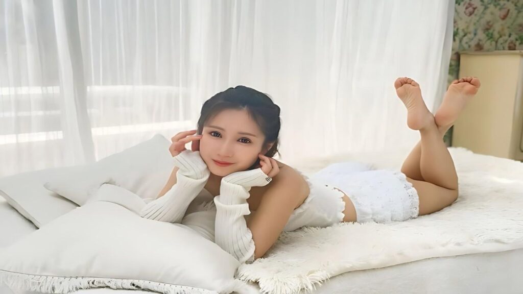 Minami Kojima lies on white bed, resting chin on her hands, smiling softly, with legs raised behind in a bright, airy room.
