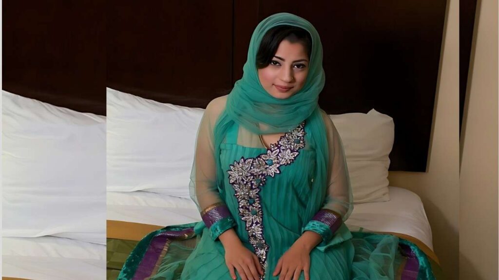 Nadia Ali in turquoise traditional dress with sheer teal scarf sits on a bed, silver-embellished panel and purple cuffs, smiling.