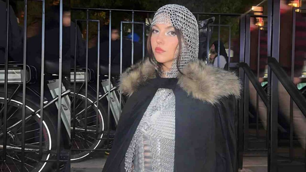 Presley Hart in metallic chainmail hood and fur-trimmed black cape, posing by a railing with bicycles behind.
