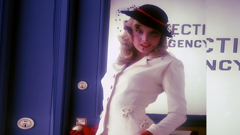 Shauna Grant in a white tailored coat with red gloves, black hat with a red band, stands by lockers beside a retro poster.