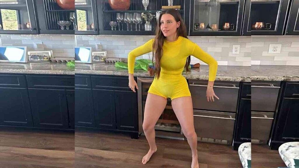 Sunny Ray in bright yellow top and matching shorts stands barefoot in a dark navy kitchen, leaning on the counter.