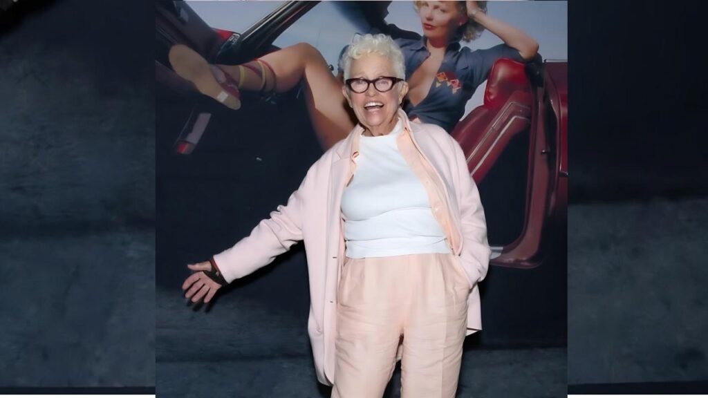 Suze Randall with short white hair and glasses, wearing a pale pink suit over a white top, arms outstretched in front of a retro car poster.