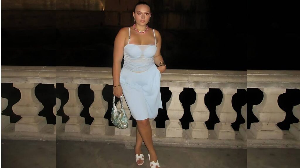 Suzi Simpson in a light blue strap dress and white sandals leans on a stone balustrade, holding a beaded bag.
