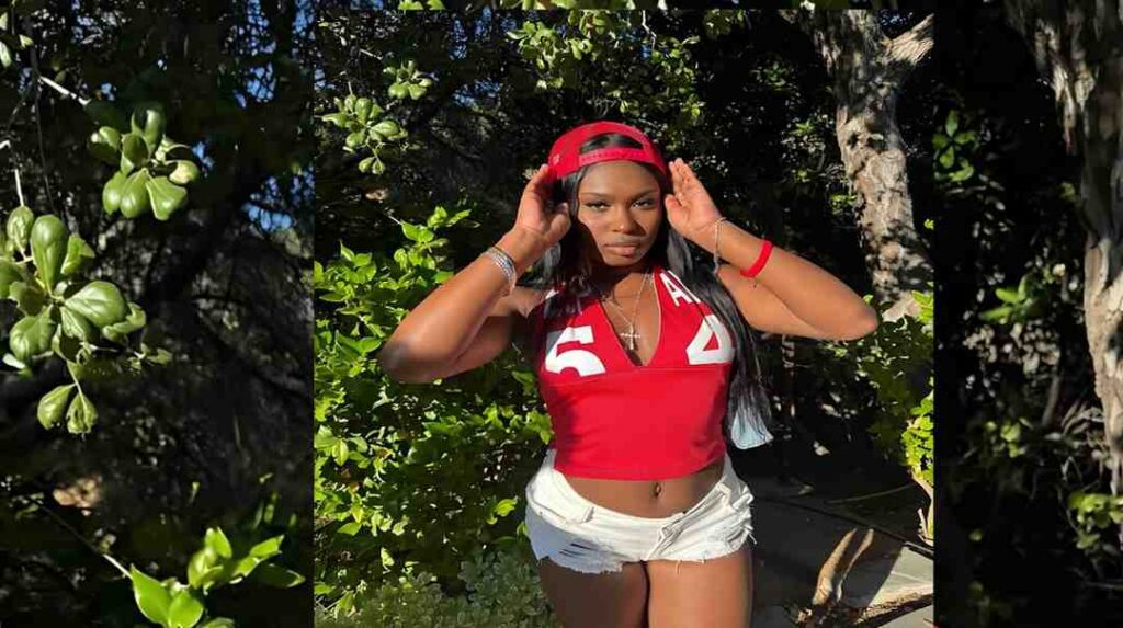 Tessa Thomas in a red cropped top and white ripped shorts, backward red cap, adjusting her cap amid greenery.