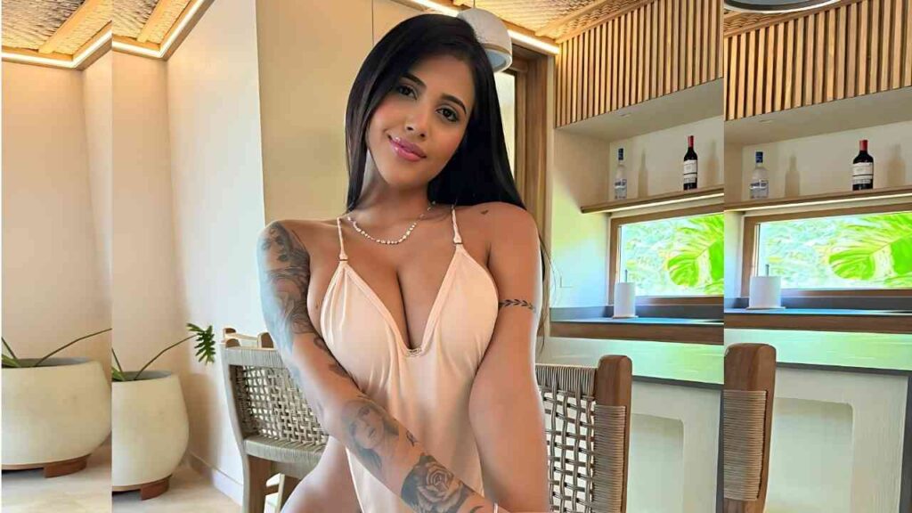 Yorgelis Carrillo with long dark hair in a pale peach slip dress sits at a wicker chair in a modern cafe, tattoos on her arms.