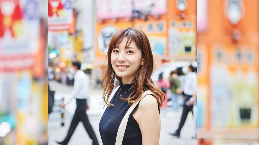 Yuma Asami in a dark sleeveless top with light straps stands in a busy street, colorful signs blurred behind her.