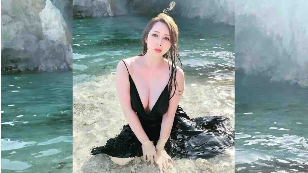Yumi Kazama (風間ゆみ ) with wet hair in a plunging black dress sits on a rock in turquoise water, waves splash around with cliffs behind.